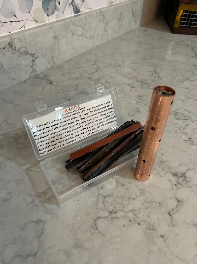 K&ouml;nnte beinhalten: Eine transparente Kunststoffbox mit Gartenschlauchstreifen und einem Kupferrohr mit L&ouml;chern. Der Text auf der Box lautet "MAGIC FIRE" und beschreibt, wie das Produkt eine farbenfrohe Lagerfeueranzeige erzeugt.