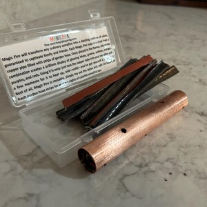 K&ouml;nnte beinhalten: Eine transparente Kunststoffbox mit einem Kupferrohr mit L&ouml;chern und mehreren Streifen Gartenschlauch. Der Text auf der Box lautet "Magic Fire" und beschreibt, wie das Produkt farbenfrohe Flammen erzeugt.