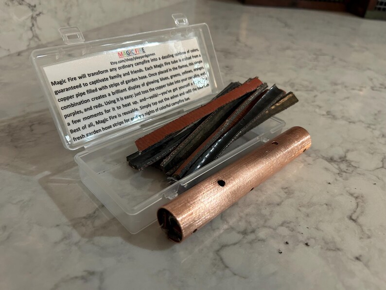 K&ouml;nnte beinhalten: Eine transparente Kunststoffbox mit einem Kupferrohr mit L&ouml;chern und mehreren Streifen Gartenschlauch. Der Text auf der Box lautet "Magic Fire" und beschreibt, wie das Produkt farbenfrohe Flammen erzeugt.