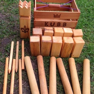 May include: A wooden Kubb game set with a wooden crate, 10 wooden blocks, a king piece, and 6 throwing sticks. The crate has a crown and the word "KUBB" engraved on it.