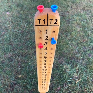 May include: A wooden scorekeeping board with two columns labeled "T1" and "T2". The board has numbers 1 through 21 marked on it. There are red and blue push pins in the holes on the board.