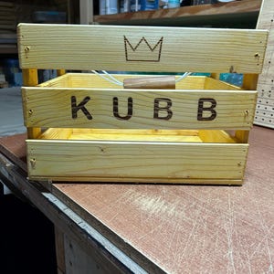 May include: A light-colored wooden crate with three horizontal slats on the front. The top slat features a laser-engraved crown icon, and the middle slat displays the word KUBB in bold, dark letters. A rope handle with a wooden grip is attached.
