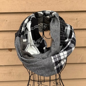 May include: A black and white infinity scarf with various patterns, including plaid and houndstooth, is displayed on a wire mannequin. The scarf is made of soft fabric and is designed to be worn around the neck.