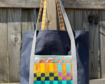 May include: A dark gray canvas tote bag with a colorful patchwork pocket. The bag has yellow and white striped straps.