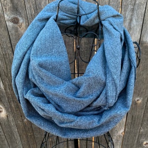 May include: A blue infinity scarf with a herringbone pattern is displayed on a black metal stand. The scarf is made of a soft fabric and is wrapped around the stand in a circular shape. The background is a weathered wooden fence.