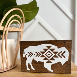 May include: A rectangular wooden sign with a dark brown stain features a white bison silhouette with a geometric design. The bison is centered on the sign, and the design includes arrows and diamond shapes. The sign is displayed on a wooden surface.