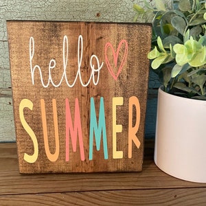 Pu&ograve; includere: Un cartello di legno con il testo "hello summer" in scrittura bianca con un cuore rosa. La parola "summer" &egrave; in lettere colorate con giallo, arancione, rosa, turchese e giallo.