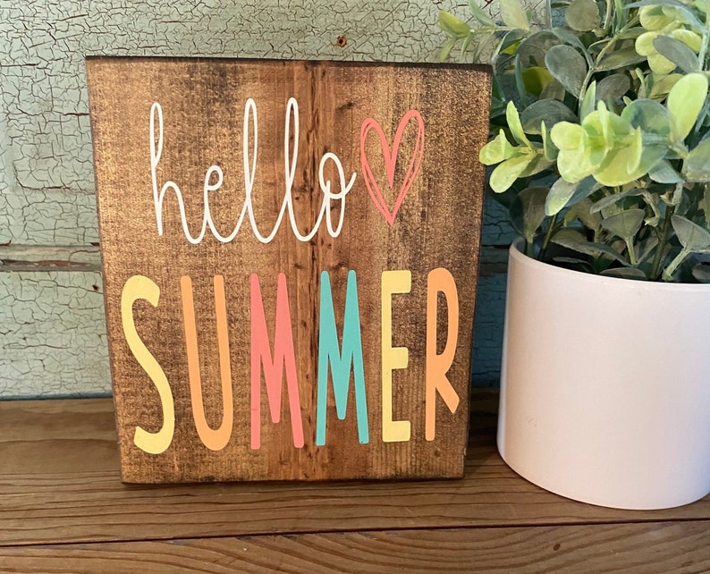 Pu&ograve; includere: Un cartello di legno con il testo "hello summer" in scrittura bianca con un cuore rosa. La parola "summer" &egrave; in lettere colorate con giallo, arancione, rosa, turchese e giallo.