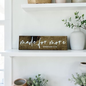 May include: A rectangular wooden sign with the phrase "made for more" in white script. The text "Ephesians 2:10" is below the main text. The sign is brown and sits on a white shelf.