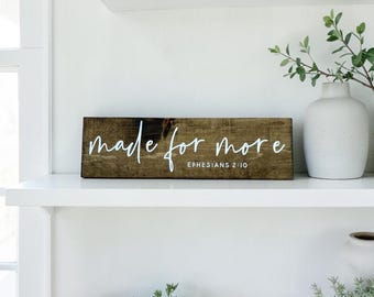 May include: A rectangular wooden sign with the phrase "made for more" in white script. The text "Ephesians 2:10" is below the main text. The sign is brown and sits on a white shelf.