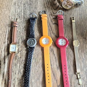 May include: A collection of wristwatches with various designs and colors. The watches include a gold-toned watch with a metal band, a brown leather strap watch with a rectangular face, and watches with colorful bands in orange and red. The watch bands are made of leather and metal.