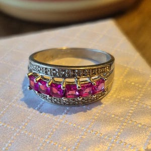 May include: Silver ring featuring a row of square, bright pink gemstones. The ring has a band with small, clear stones. The ring is set on a white surface.