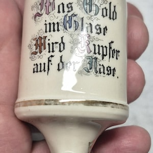 May include: A white ceramic bottle stopper with a gold rim and a spout. The bottle stopper has a metal lid with a handle. The bottle stopper has a gold inscription that reads "Was Gold im Glasse Wird Kupfer auf der Nase."