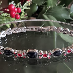 May include: Silver-toned bracelet with black and red accents. The bracelet features a series of black, oval-shaped stones and red, round stones set in a silver metal band. The clasp is a fold-over style.