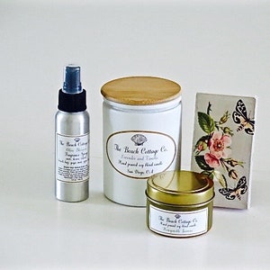 May include: Three products from The Beach Cottage Co. in a white setting. A silver spray bottle with a black cap and a label that reads "The Beach Cottage Co. Olive Blossom Fragrance Spray". A white candle jar with a wooden lid and a label that reads "The Beach Cottage Co. Lavender and Tahitian Hand poured soy blend candles New Smyrna, FL". A small gold tin candle with a label that reads "The Beach Cottage Co. Hand poured soy blend candles Honeycomb & Tahitian".