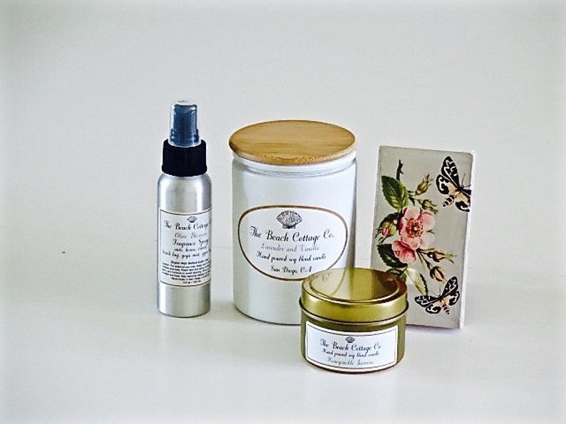 May include: Three products from The Beach Cottage Co. in a white setting. A silver spray bottle with a black cap and a label that reads "The Beach Cottage Co. Olive Blossom Fragrance Spray". A white candle jar with a wooden lid and a label that reads "The Beach Cottage Co. Lavender and Tahitian Hand poured soy blend candles New Smyrna, FL". A small gold tin candle with a label that reads "The Beach Cottage Co. Hand poured soy blend candles Honeycomb & Tahitian".