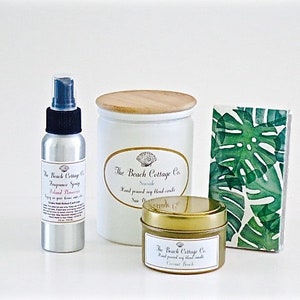 May include: A set of three bath and body products from The Beach Cottage Co. The set includes a white candle with a wooden lid, a gold tin candle, and a silver spray bottle. The candle tins have a white label with the company name and product description. The spray bottle has a white label with the company name, product description, and the scent "Island Paradise". The set is displayed on a white surface.