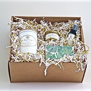 May include: A brown cardboard box filled with white and gold shredded paper. The box contains a white candle jar with a gold lid, a small gold jar with a gold lid, a small clear glass bottle with a gold lid, and a green and white patterned soap bar.