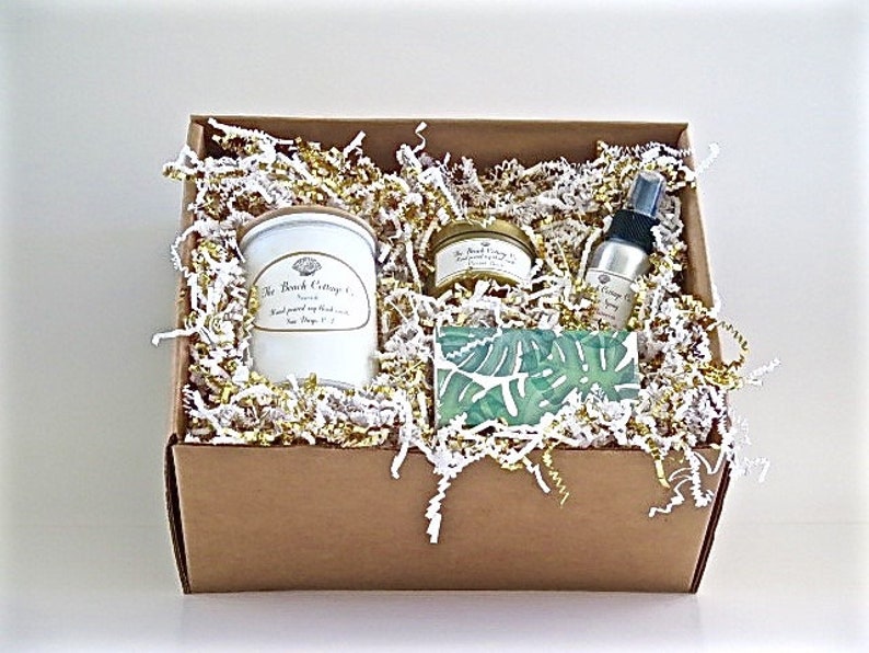 May include: A brown cardboard box filled with white and gold shredded paper. The box contains a white candle jar with a gold lid, a small gold jar with a gold lid, a small clear glass bottle with a gold lid, and a green and white patterned soap bar.