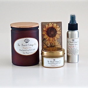 May include: A set of three fall-themed home fragrance products from The Beach Cottage Co.  The set includes a brown glass candle with a wooden lid labeled "Pumpkin Spice", a small gold tin candle labeled "Caramel Rice", and a silver spray bottle labeled "Sandalwood Rose".