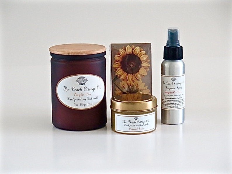 May include: A set of three fall-themed home fragrance products from The Beach Cottage Co.  The set includes a brown glass candle with a wooden lid labeled "Pumpkin Spice", a small gold tin candle labeled "Caramel Rice", and a silver spray bottle labeled "Sandalwood Rose".