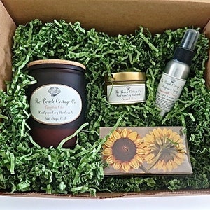 May include: A gift box filled with green shredded paper and a brown candle with a wooden lid, a small gold jar with a lid, and a silver spray bottle. The candle and jar have labels that say "The Beach Cottage Co." The spray bottle has a label that says "The Beach Cottage Co. Fragrance Spray". The box also contains a card with a sunflower design.