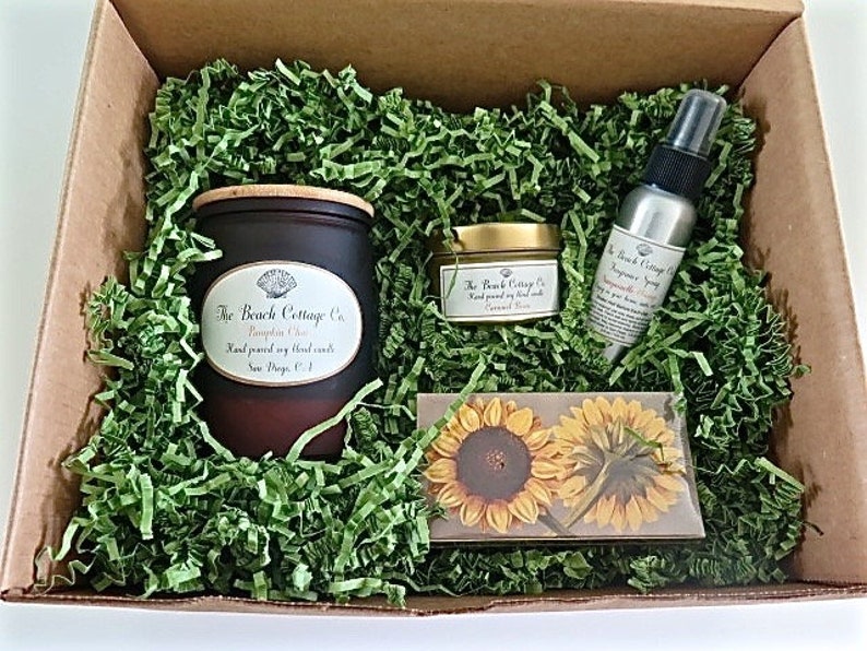 May include: A gift box filled with green shredded paper and a brown candle with a wooden lid, a small gold jar with a lid, and a silver spray bottle. The candle and jar have labels that say "The Beach Cottage Co." The spray bottle has a label that says "The Beach Cottage Co. Fragrance Spray". The box also contains a card with a sunflower design.