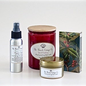 May include: A set of three candles in glass jars with wooden lids. The candles are labeled "The Beach Cottage Co." and have the scents "Spiced Cranberry", "Frosted Sugarplum", and "Cranberry Spice". The candles are packaged in a box with a holly design.