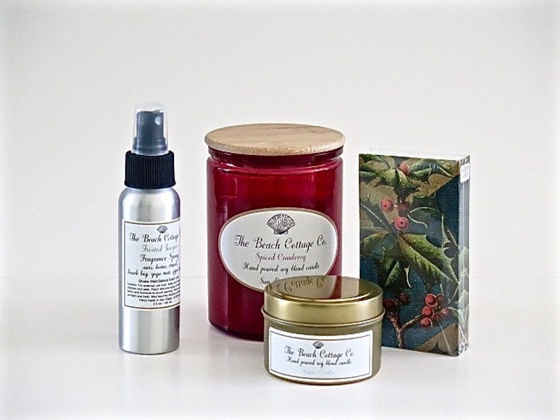 May include: A set of three candles in glass jars with wooden lids. The candles are labeled "The Beach Cottage Co." and have the scents "Spiced Cranberry", "Frosted Sugarplum", and "Cranberry Spice". The candles are packaged in a box with a holly design.
