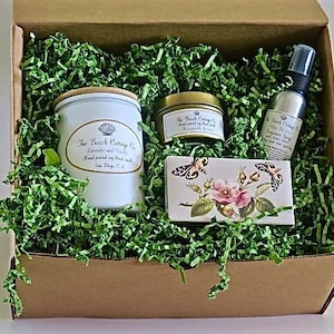 May include: A gift box filled with green shredded paper and a variety of bath and body products. The products include a candle, a jar of body butter, and a bottle of room spray. The products are all labeled with the text "The Beach Cottage Co."