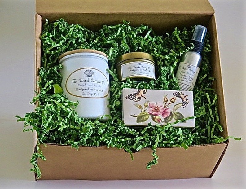May include: A gift box filled with green shredded paper and a variety of bath and body products. The products include a candle, a jar of body butter, and a bottle of room spray. The products are all labeled with the text "The Beach Cottage Co."