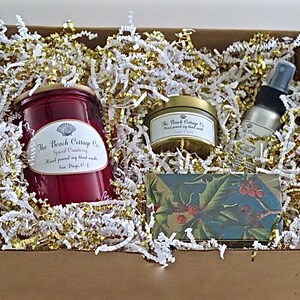 May include: A gift box filled with white and gold shredded paper. The box contains a red candle with a white label that reads "The Beach Cottage Co. Spiced Cranberry Hand poured soy blend candle San Diego, CA", a small gold tin candle with a white label that reads "The Beach Cottage Co. Hand poured soy blend candle", a small bottle of room spray with a white label that reads "The Beach Cottage Co. Spiced Cranberry", and a small rectangular box with a green and red holly design.