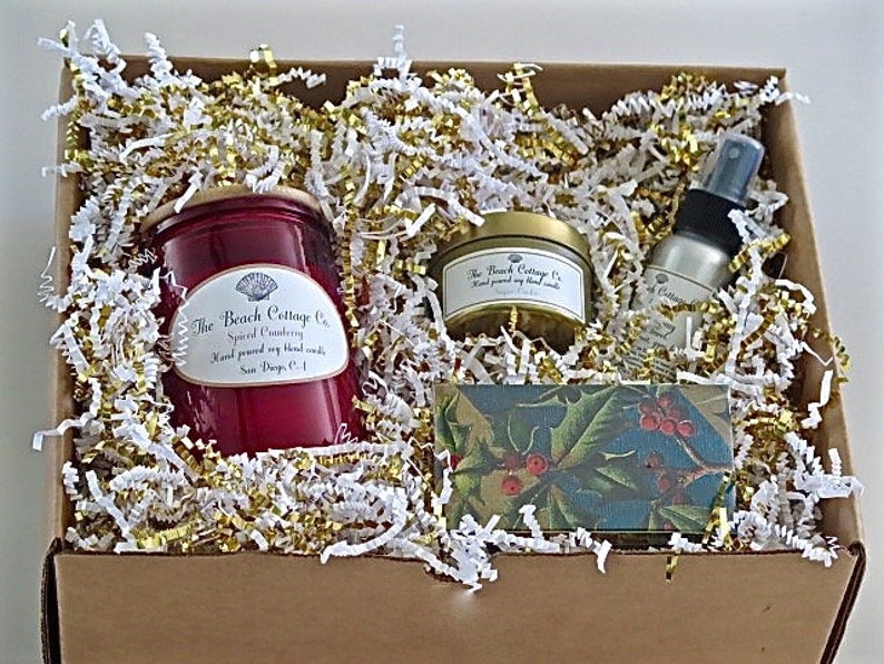 May include: A gift box filled with white and gold shredded paper. The box contains a red candle with a white label that reads "The Beach Cottage Co. Spiced Cranberry Hand poured soy blend candle San Diego, CA", a small gold tin candle with a white label that reads "The Beach Cottage Co. Hand poured soy blend candle", a small bottle of room spray with a white label that reads "The Beach Cottage Co. Spiced Cranberry", and a small rectangular box with a green and red holly design.