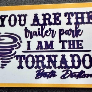 May include: A rectangular sign with a white background and a yellow border. The sign features the text "YOU ARE THE trailer park I AM THE TORNADO Both Dutton" in dark blue lettering, with a stylized tornado graphic.