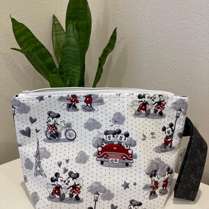 May include: White zippered pouch with a black and white print of Mickey Mouse and Minnie Mouse in various scenes. The print includes hearts, stars, a red car, and the Eiffel Tower. The pouch has a black zipper and a black strap.