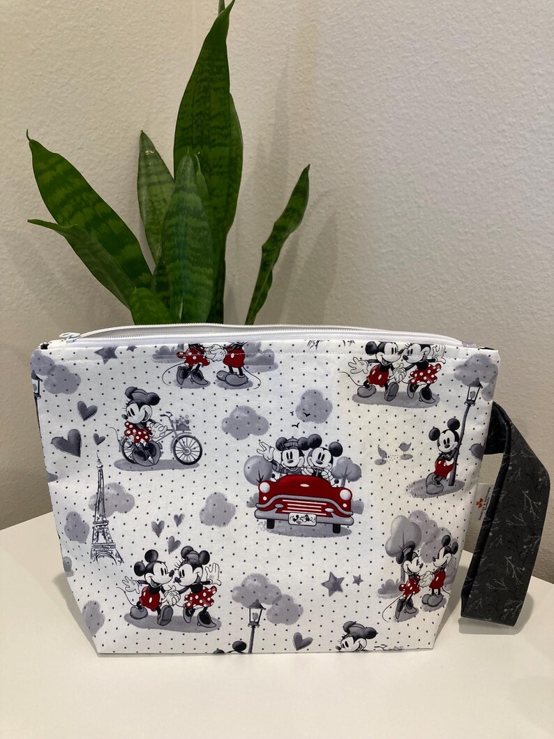 May include: White zippered pouch with a black and white print of Mickey Mouse and Minnie Mouse in various scenes. The print includes hearts, stars, a red car, and the Eiffel Tower. The pouch has a black zipper and a black strap.