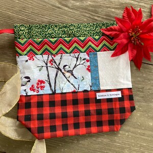 May include: A red and black plaid drawstring bag with a white fabric panel featuring a wintery scene with birds and berries. The bag has a green and red chevron pattern and a green swirl pattern on the top. The bag has a tag that says "Sunshine & Bubblegum".