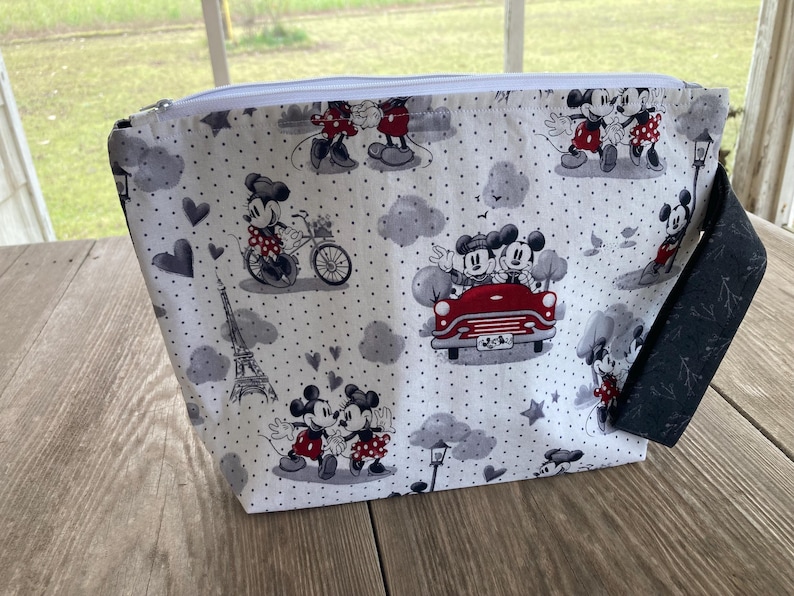 May include: A white zippered pouch with a black and white polka dot pattern featuring Mickey Mouse and Minnie Mouse in various scenes. The pouch has a black zipper and a black strap.