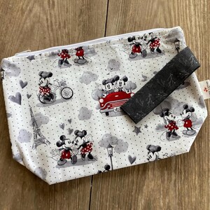 May include: A white zippered pouch with a black and white polka dot pattern. The pouch features a repeating pattern of Mickey Mouse and Minnie Mouse in various poses, including riding a bicycle, driving a car, and holding hands. The pouch has a black zipper and a black fabric handle.
