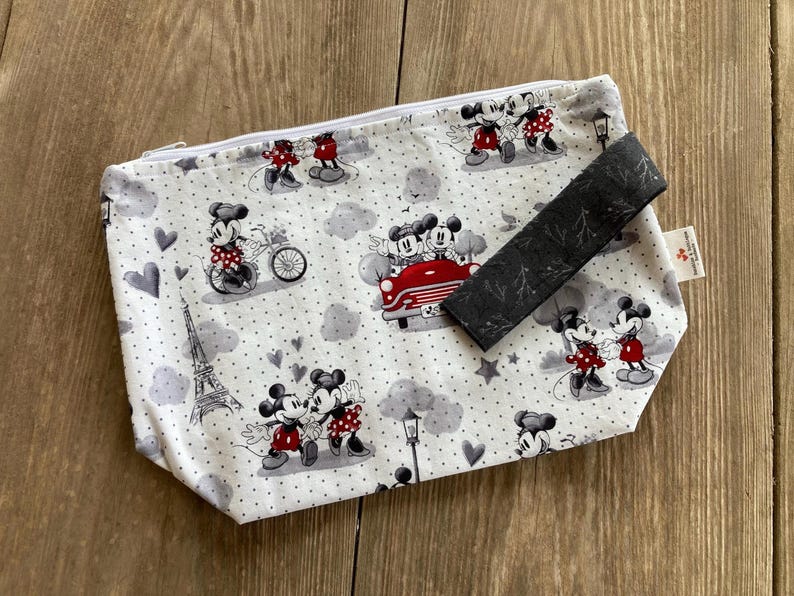 May include: A white zippered pouch with a black and white polka dot pattern. The pouch features a repeating pattern of Mickey Mouse and Minnie Mouse in various poses, including riding a bicycle, driving a car, and holding hands. The pouch has a black zipper and a black fabric handle.