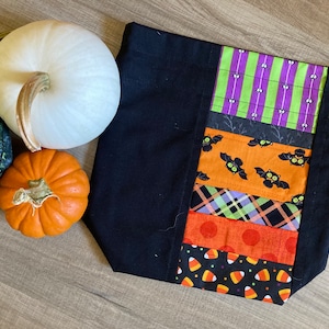 Op de afbeelding: Een zwarte stoffen trekkoordtas met een Halloween-thema. De tas is versierd met kleurrijke stofstroken met vleermuizen, snoepmais en stippen.