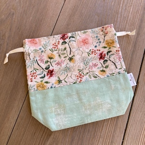 May include: A drawstring bag with a floral pattern on the top half and a solid light green bottom. The floral pattern includes pink, red, and yellow flowers with green leaves on a cream background. The bag is made of fabric.