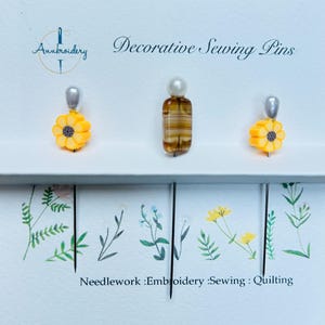 May include: Decorative sewing pins with floral and pearl designs. One pin features a yellow flower with a silver top, another has a pearl top, and the third has a striped amber design. The text reads "Decorative Sewing Pins".
