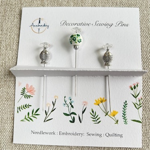 May include: A set of decorative sewing pins presented on a white card. The pins feature distinctive toppers, such as a fish and a floral pattern. The card is printed with "Decorative Sewing Pins" and "Needlework: Embroidery: Sewing: Quilting."