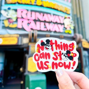 May include: A white sticker with a red border and the text "Nothing can stop us now!" in red. Mickey Mouse and Minnie Mouse are on the sticker, with Mickey Mouse on the left and Minnie Mouse on the right. The sticker is being held in front of a building with a sign that says "Mickey & Minnie's Runaway Railway".
