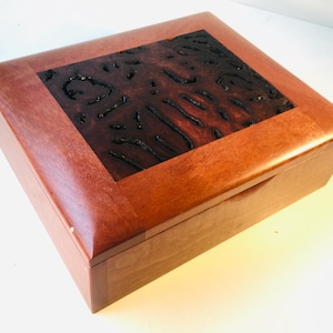 May include: A rectangular wooden box in a reddish-brown hue. The lid incorporates a dark, textured inlay with a distinctive pattern. The box is designed with a simple, elegant aesthetic, suitable for storage or display.