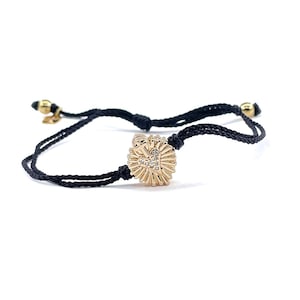 May include: A black cord bracelet with a gold charm featuring a heart with a diamond-like design. The charm is surrounded by a sunburst pattern.