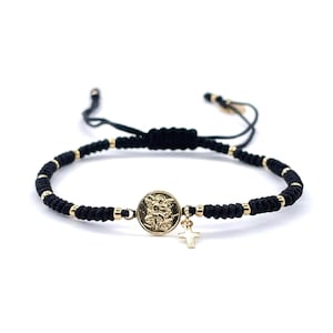 May include: A black string bracelet with gold beads and a gold charm featuring a religious icon. The charm is attached to a small gold cross.