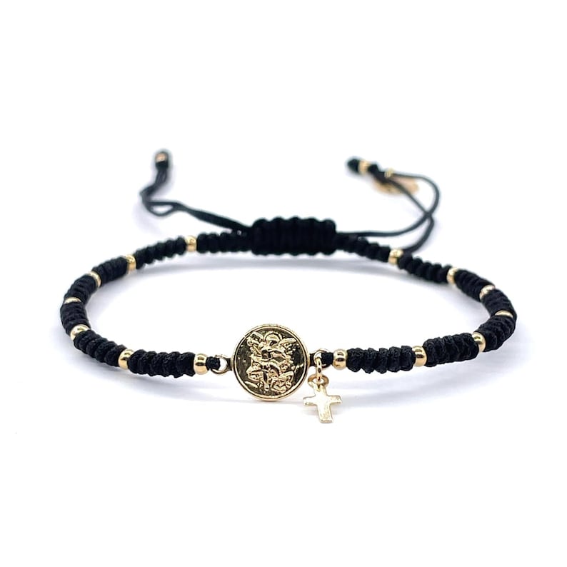 May include: A black string bracelet with gold beads and a gold charm featuring a religious icon. The charm is attached to a small gold cross.