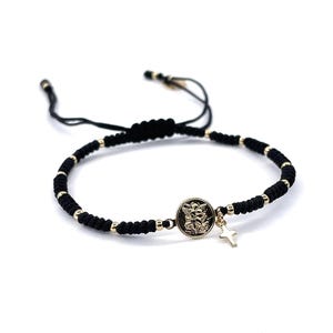 May include: A black string bracelet with gold beads and a gold charm featuring a religious image. The charm is attached to a small gold cross.